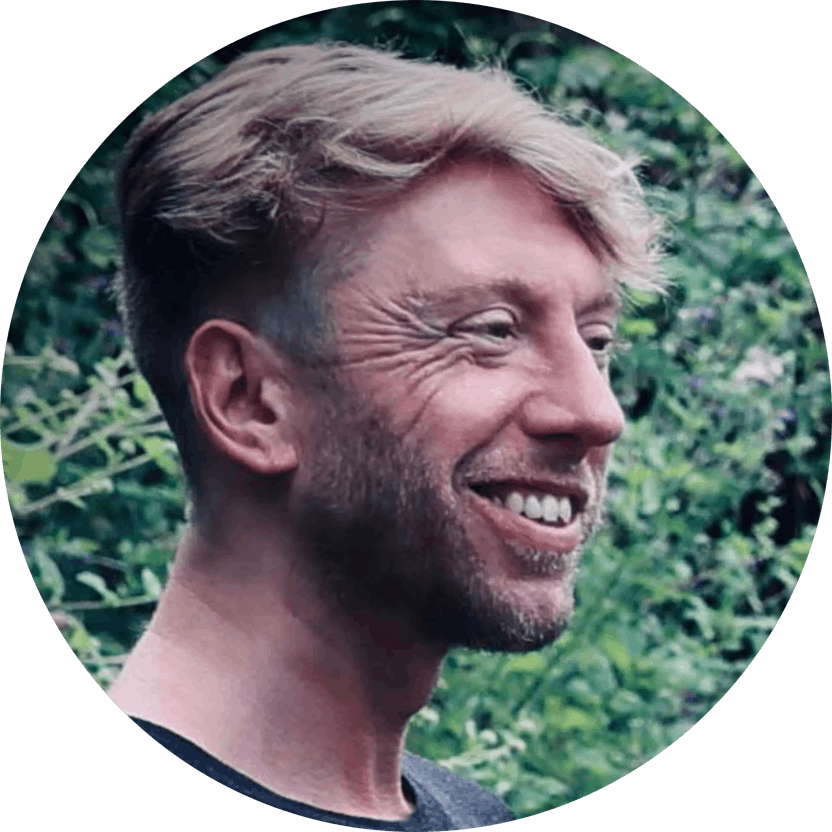 a picture of Jamie C Goodwin a male watercolour artist born in 1985. He is outside in the forest of his off-grid home, looking to the right, with a cheaky grin. It's day time, and the sun is warm from the summer.