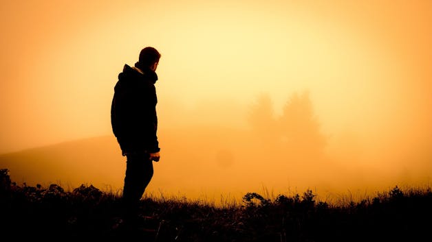 Silhouette of a man walking toward a glowing sunrise in a misty field, evoking a journey of faith.