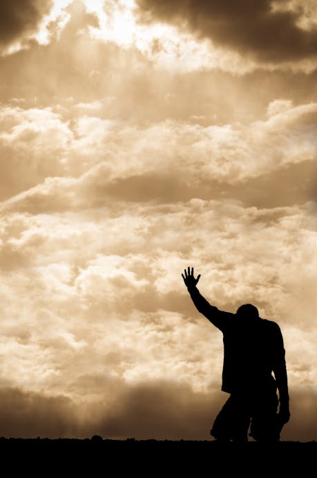 Silhouette of a man kneeling with one hand raised toward light breaking through the clouds, symbolizing calling, faith, and dedication.