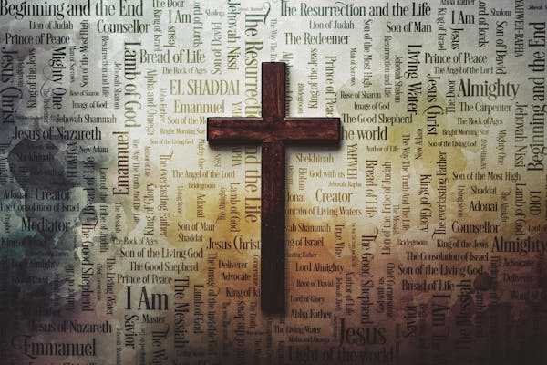 A wooden cross centrally positioned over a background filled with various names and titles attributed to Jesus Christ, written in different fonts and sizes, creating a mosaic of words from Christian theology.