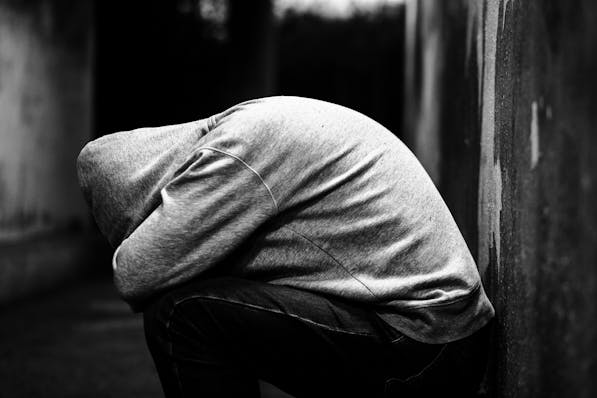 A monochrome image capturing the poignant moment of a person crouching down with their head bowed, concealed by a hoodie, in a dimly lit environment that evokes a sense of despair or contemplation.