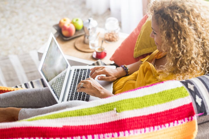 Colourful home scene of a woman lying on the sofa while working on her laptop